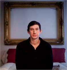 a man sitting on a bed in front of a framed photo above his head, looking at the camera