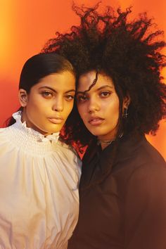 two women standing next to each other in front of an orange wall