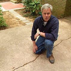 an older man squatting down on the ground in front of a brick building with trees and bushes behind him