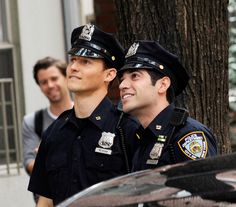 two police officers standing next to each other in front of a tree and people on the sidewalk