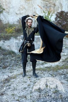 a man dressed in black and gold holds his cape over his head as he stands on a rocky area