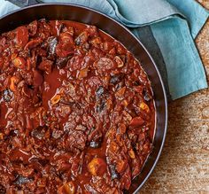 a pan filled with chili and meat on top of a wooden table next to a blue napkin