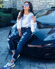 a woman sitting on top of a black sports car wearing blue jeans and white shirt