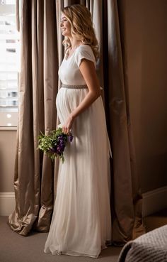 a pregnant woman in a white dress standing next to a window holding a bouquet of flowers