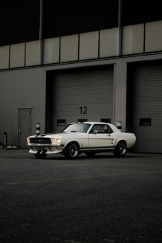 a white muscle car parked in front of a building with two garage doors on it