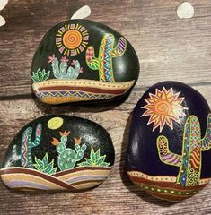 three painted rocks sitting on top of a wooden table next to flowers and cacti