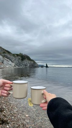 two people holding coffee mugs in their hands on the shore of a body of water