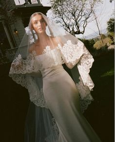 a woman wearing a wedding dress and veil in front of a house with trees on the other side