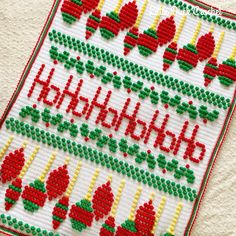 a cross stitch christmas card with red and green ornaments on it's side, sitting on a white surface