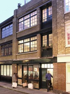 two people are walking down the sidewalk in front of a building with many windows and doors