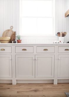 a kitchen with white cabinets and wood floors in front of a window that has potted plants on top of it