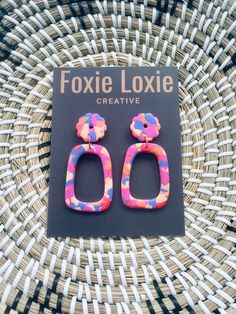 a pair of pink and blue earrings sitting on top of a woven basket next to a card