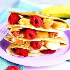 three pancakes with bananas and raspberries on a purple plate next to a banana