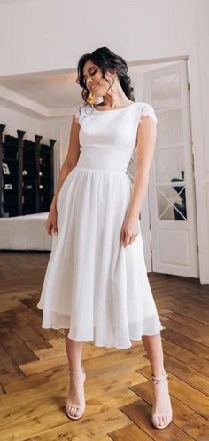 a woman standing on top of a wooden floor wearing a white dress and sandals with her hand in her mouth