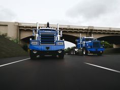 two large blue trucks driving down a street next to a bridge and road overpass