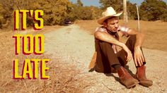 a man sitting on the side of a dirt road wearing a cowboy hat and boots