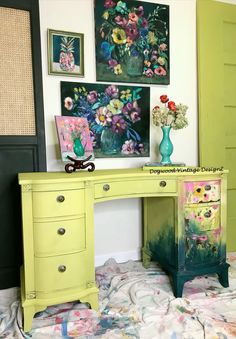 a green desk with flowers on it in front of pictures and paintings hanging on the wall