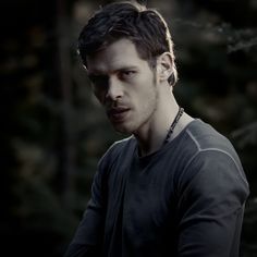 a man in black shirt standing next to trees and looking at the camera with serious look on his face