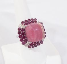 "A sublime art deco design ring, celebrating the marriage of a one-of-a-kind rhodochrosite gemstone, framed at its base by 4 carats of rich rose-red Rhodolite garnets in a double halo. Accented further by a cinematic setting of sterling silver at the far four corners of the center stone. This 15mm cushion cut natural rhodochrosite is a high-profile organic beauty. The ivory toned banding on one side is a wonderful contrast to the luscious strawberry color of this unique stone. From any angle, wh Rhodalite Garnet Jewelry, Round Rhodonite Jewelry With Natural Stones, Rhodochrosite Jewelry Rings, Pink Aragonite, Rhodochrosite Jewelry, Strawberry Color, Double Halo, Rhodolite Garnet, Organic Beauty