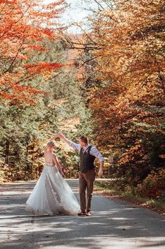 a bride and groom are walking down the road in front of trees with orange leaves