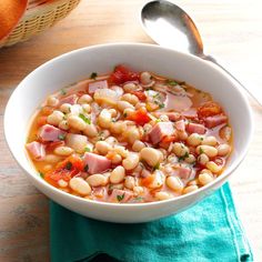 a white bowl filled with beans and ham on top of a green napkin next to a spoon