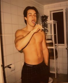 a shirtless man brushing his teeth in the bathroom with an open window behind him