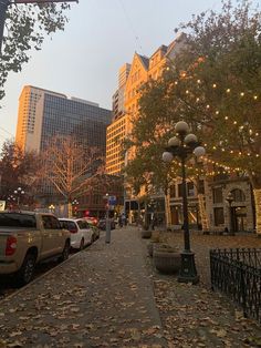 a city street lined with parked cars next to tall buildings covered in fall leaves and lights