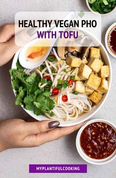 two hands holding a bowl of food with tofu and sauces on the side