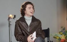 a young man sitting in a chair laughing and holding a piece of paper while wearing a brown leather jacket