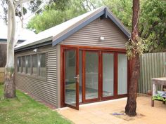 a small shed with sliding glass doors on the front and side walls, sitting next to a tree