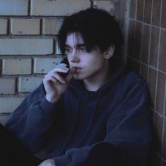 a young man is talking on his cell phone while leaning up against a brick wall