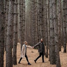 a man and woman holding hands walking through a forest filled with tall pine trees on a sunny day