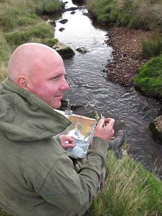 a man sitting in the grass next to a stream holding a metal container with something on it