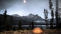 a tent pitched up on the ground in front of some trees and mountains under a cloudy sky