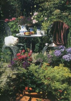 a table and chairs in a garden with lots of flowers on the ground next to it