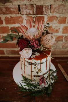 a wedding cake with flowers and greenery is on a table next to a brick wall