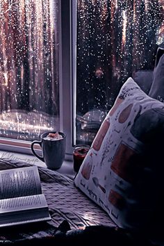 an open book on a table next to a window with rain falling down the outside