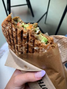 a person holding up a sandwich wrapped in brown paper with broccoli and cheese on it