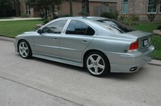 a silver car parked in front of a house on the side of a street with grass and trees behind it