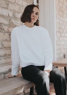 a woman sitting on top of a wooden bench next to a brick building and wearing black pants