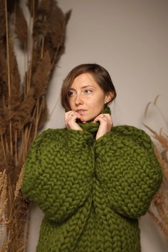 a woman in a green sweater standing next to some tall grass and dried plants with her hands on her chin