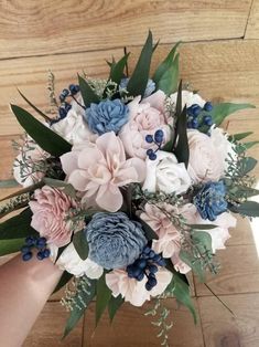 a bridal bouquet with blue and pink flowers on a wooden table next to a person's hand