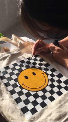 a woman is painting a smiley face on a tote bag with black and white checkered fabric