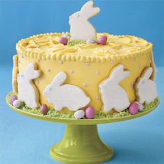 a yellow cake decorated with white rabbits and eggs on a green stand against a blue background