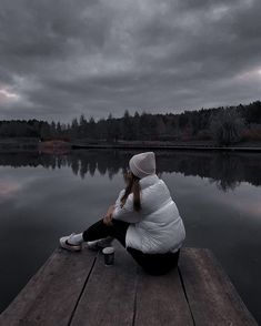 a woman sitting on a dock next to a body of water with trees in the background