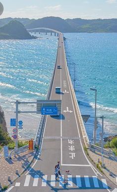 two people crossing the street on a long bridge over looking the ocean and hills in the distance