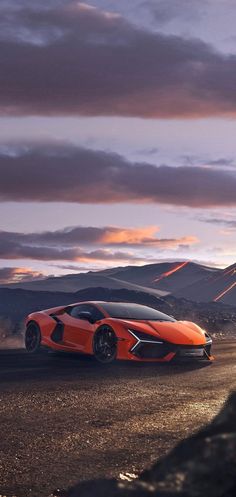 an orange sports car driving down the road at dusk with mountains in the background and cloudy skies