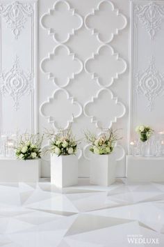 three white vases with flowers in them sitting on the floor next to a wall