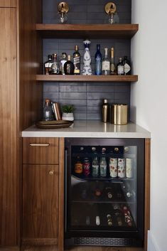 a small bar with bottles and glasses on the top shelf, next to an open wine cooler