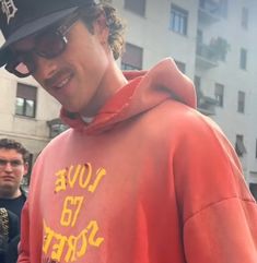 a young man wearing a red sweatshirt and black hat standing in front of a building